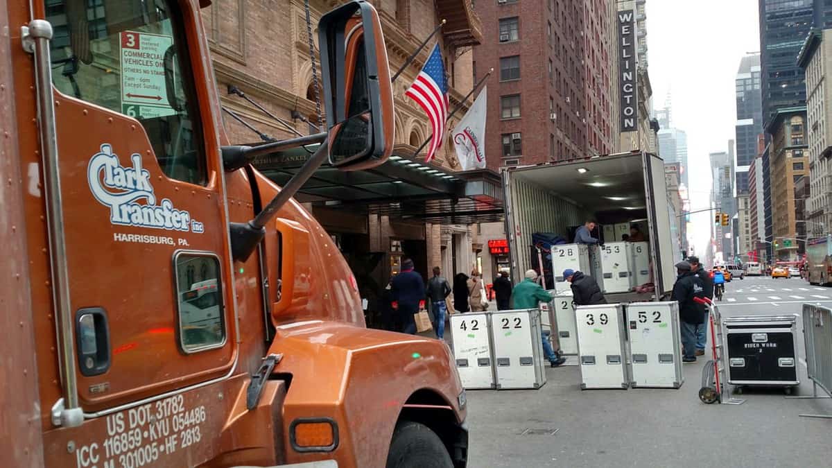 A photograph of a truck on a road lined with city buildings. In front of the truck are numbered boxes and the open trailer of another truck.