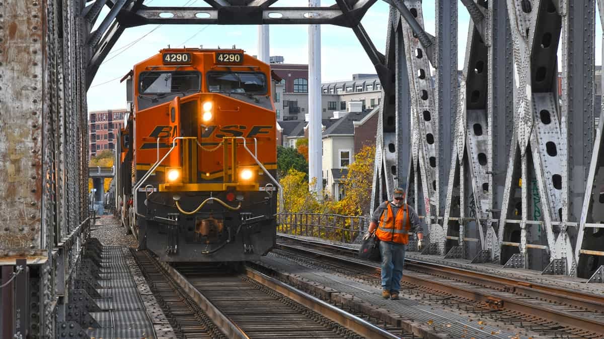 A photograph of a freight train passing through a railroad bridge. A railroad worker is standing next to the train.