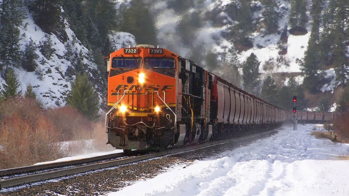 A photograph of a train traveling on rail track. There is snow on the ground.