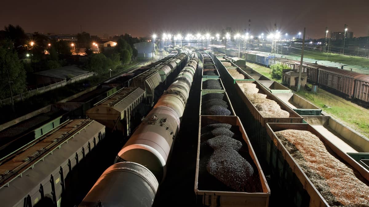 A photograph of a rail yard at night. Inside the rail yard are eight long rows of parked railcars.