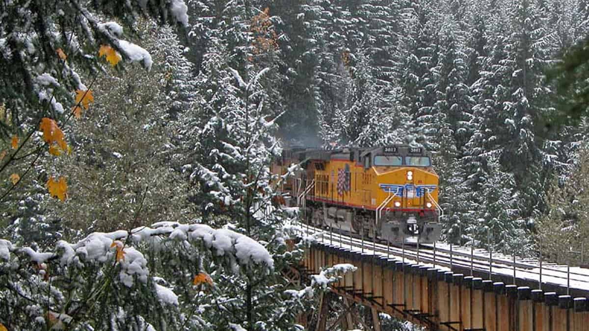 A photograph of a train that is about to get onto a rail bridge. There is snow on the trees by the bridge.