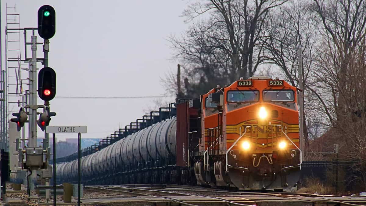 A photograph of a train with its headlights on. The day is cloudy. The locomotive is hauling a line of tank cars behind it.