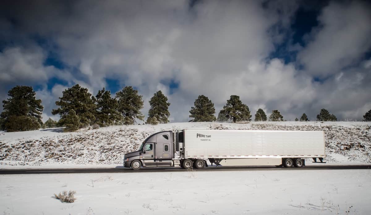 Tractor-trailer moving down a road with snowy hills in the background.