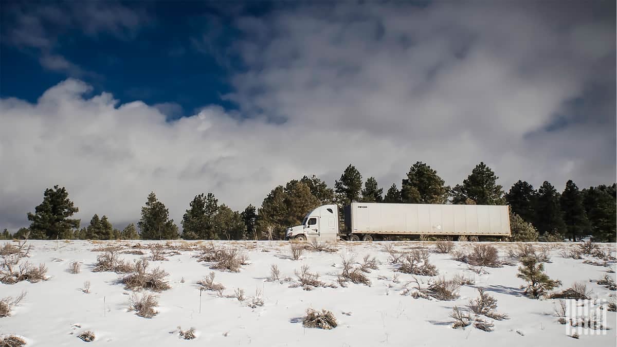 Tractor-trailer moving down snowy road and hillside.