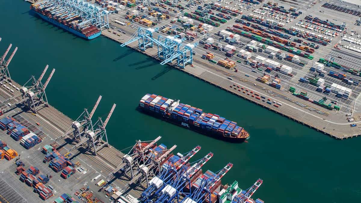 A photograph of a container vessel sailing into a shipping channel at a port. There are cranes at the dock and hundreds of container boxes sitting on land.