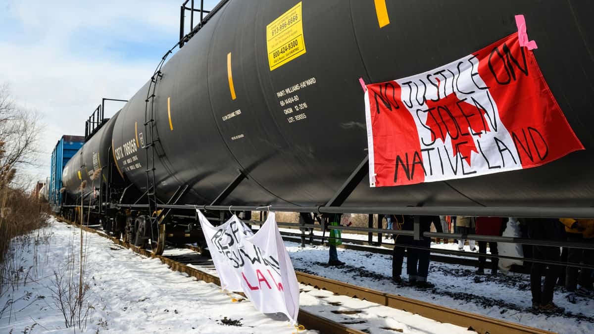 A photograph of tank cars on a rail track. One of the tank cars has a Canadian flag hanging from it. Written on the flag are the words, "no justice on stolen native land."