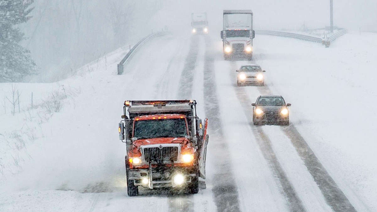 Plow clearing snowy Michigan road, with cars and a tractor-trailer behind it.