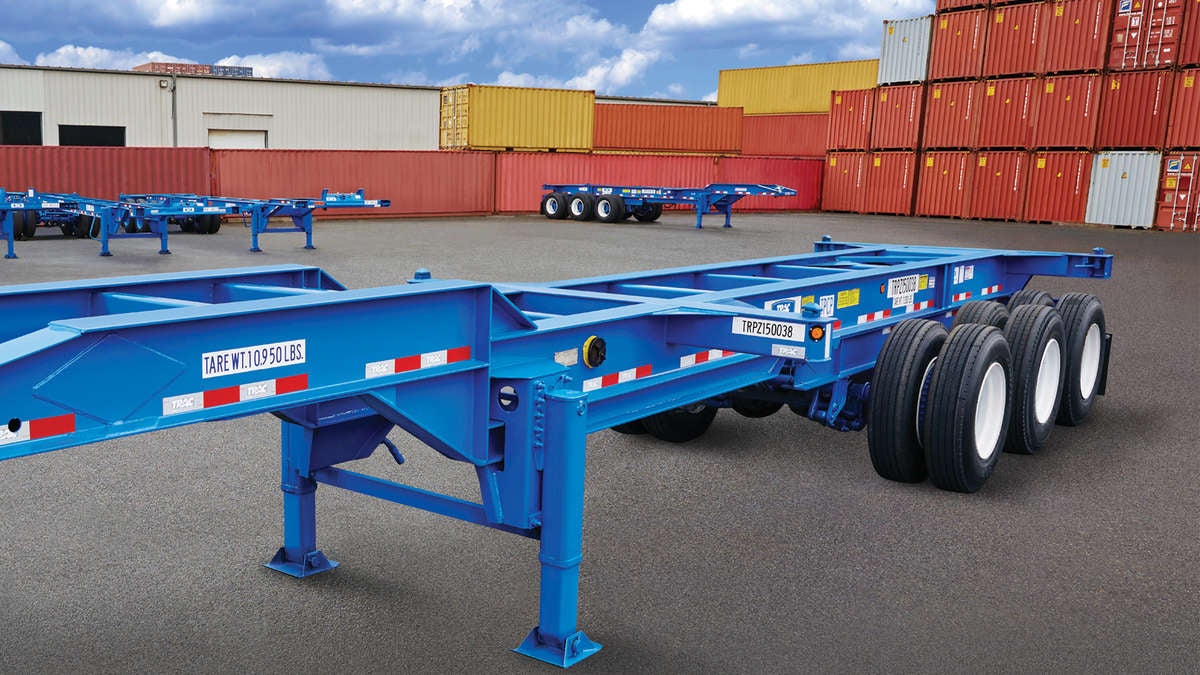 A photograph of an intermodal chassis at a container yard. There are intermodal containers stacked in rows behind the chassis.