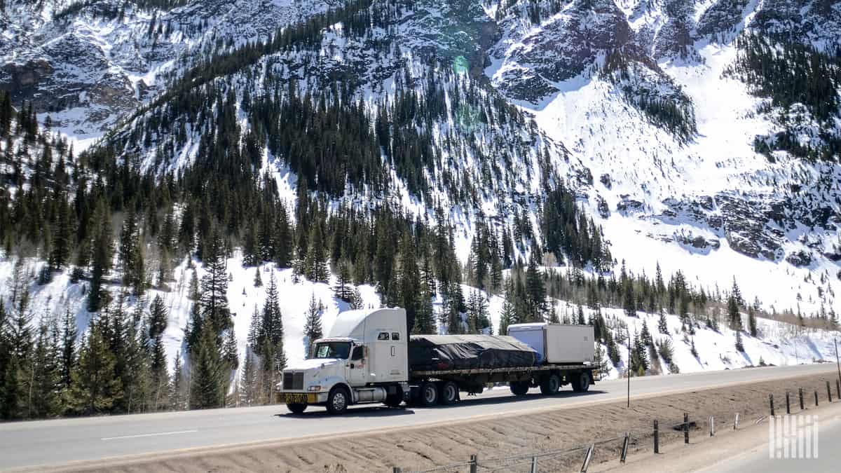 Tractor-trailer on mountain road with snow in background.