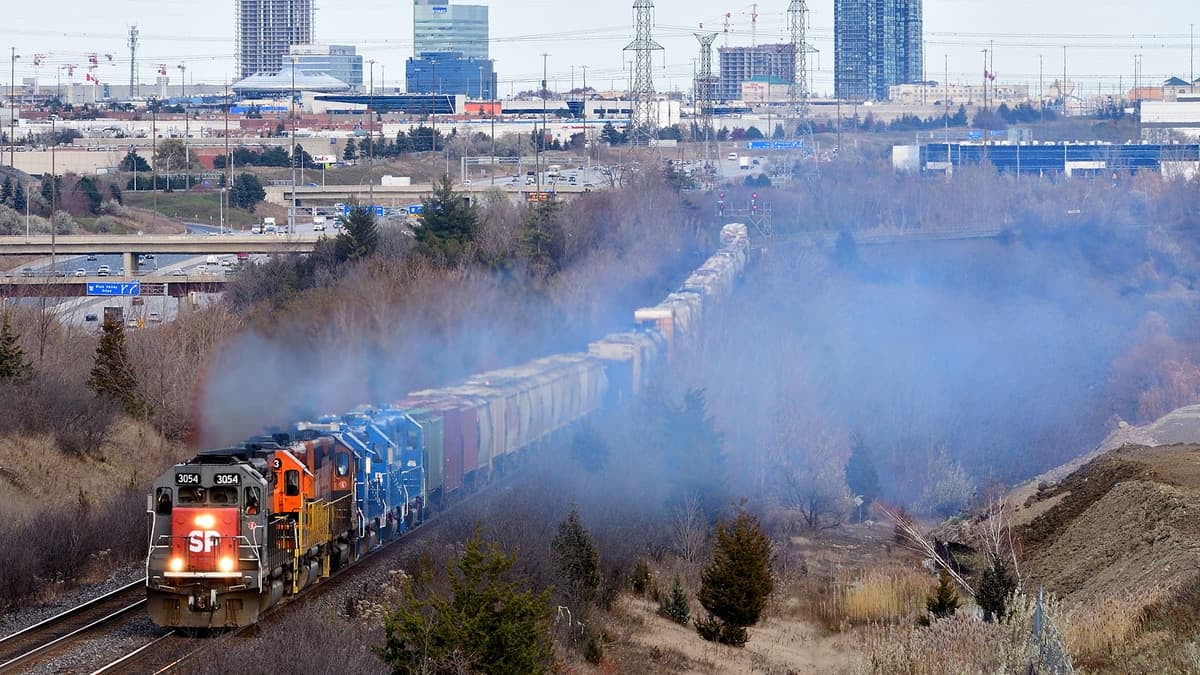 A photograph of a train traveling through a wooded area. There are city buildings on the horizon.