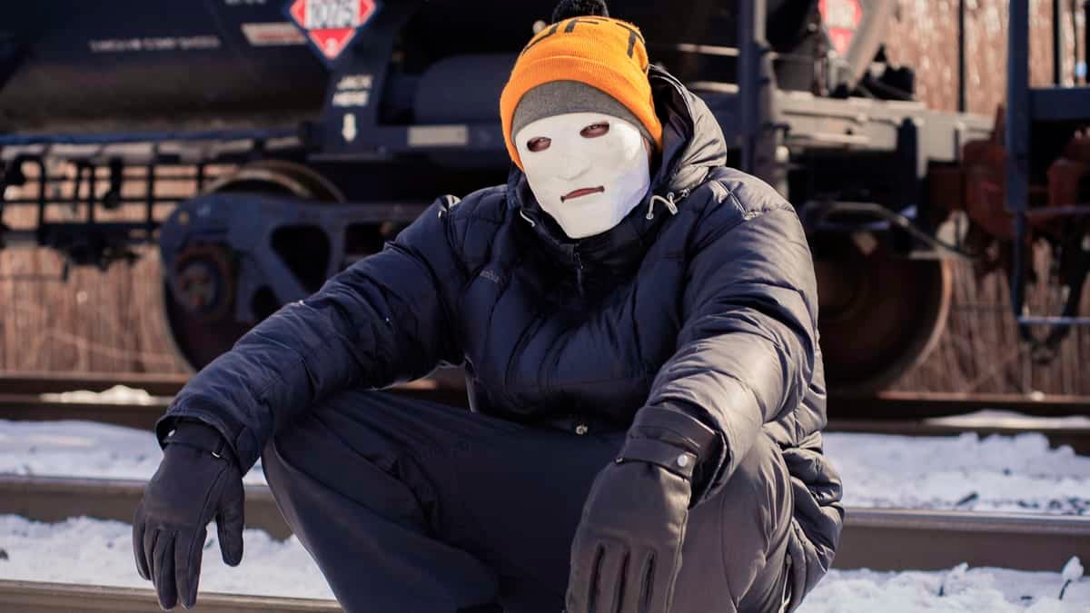 A photograph of a man with a mask on his face. He is sitting next to a train.