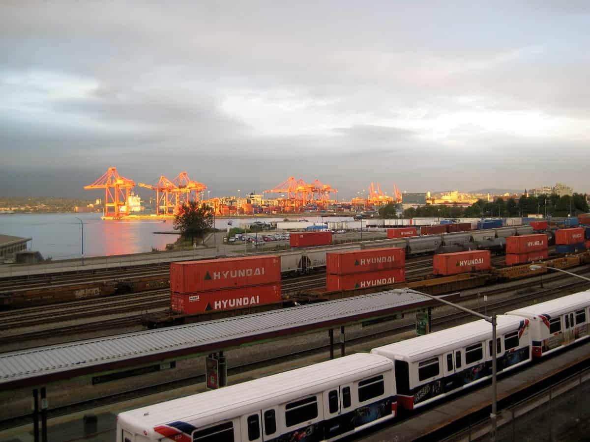 A photograph of a rail yard. There are intermodal containers and rail cars There are loading cranes at a port in the background.