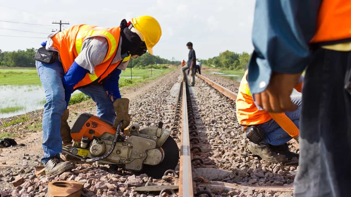 A photograph of two workers fixing a railroad track. A third person is standing on the railroad track in the distance.