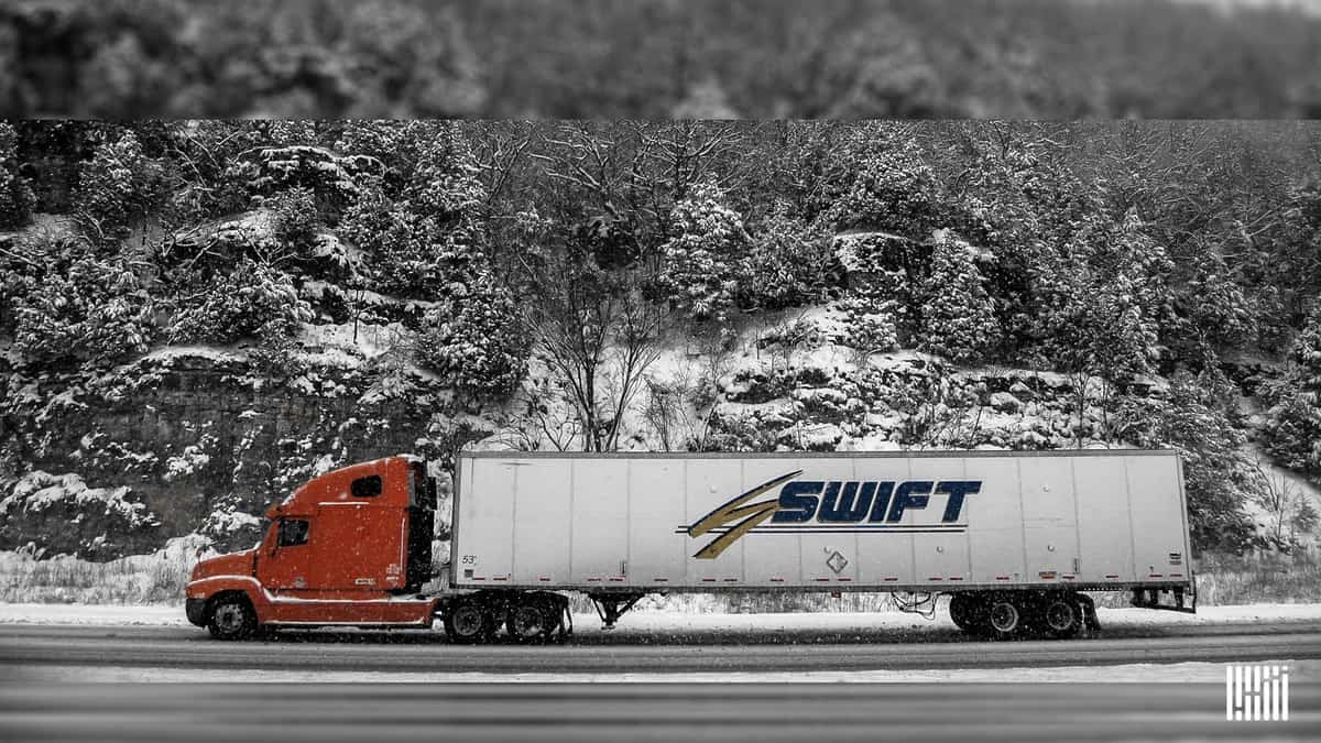 Tractor-trailer heading down snow road with snowy hillside in background.