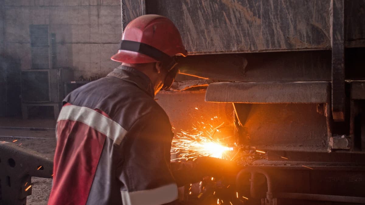 A photograph of a man. His back is to the viewer. The worker is repairing equipment and there is a fire near the worker.