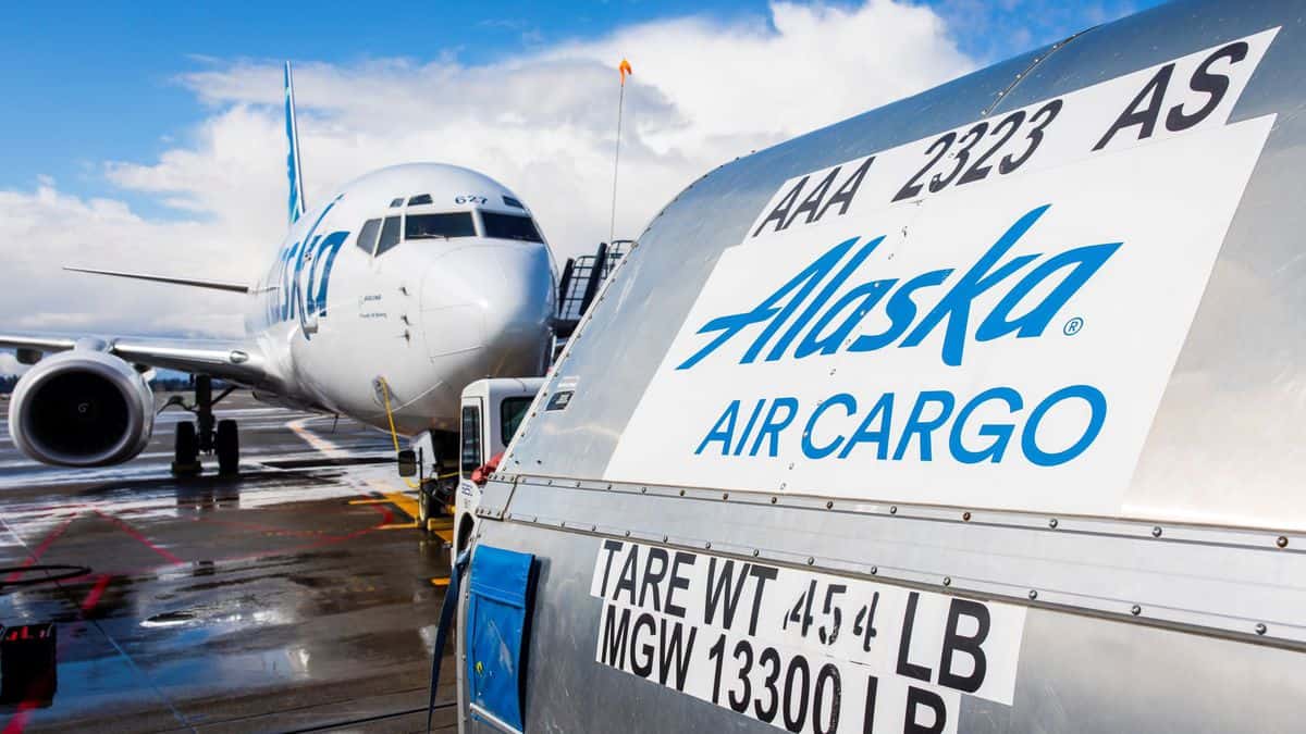 An Alaska Airlines shipping container sits on tarmac in front of plane.