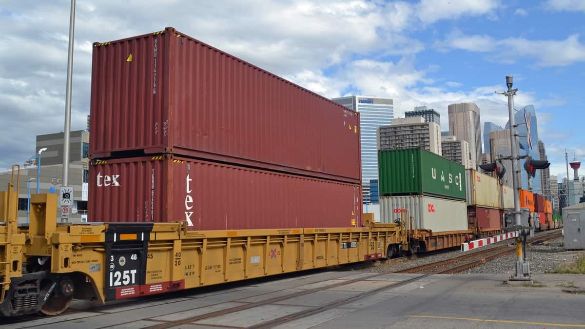 A photograph a train carrying intermodal containers. Tall city buildings are in the background.