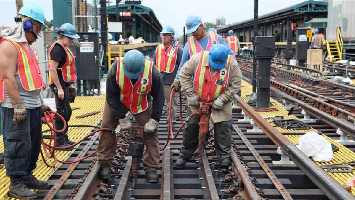 A photograph of workers performing work on a rail track near a passenger railway station.