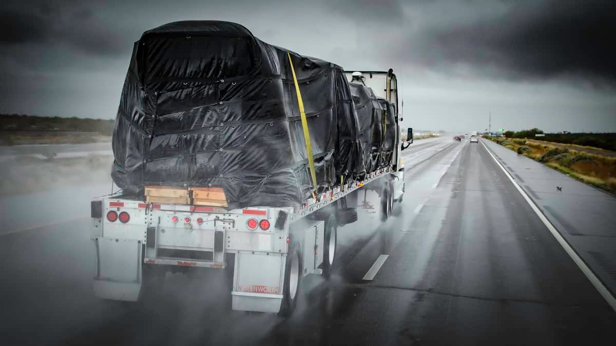 Covered flatbed tractor-trailer on wet highway.