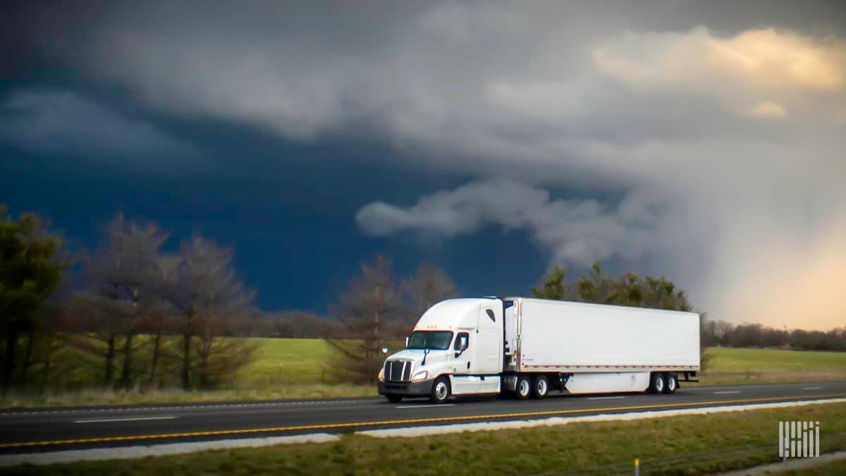 Tractor-trailer heading down highway with thunderstorm cloud in background.