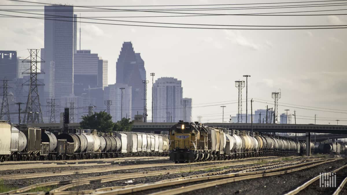 A photograph of a train at a rail yard. There are tall buildings and skyscrapers behind it.