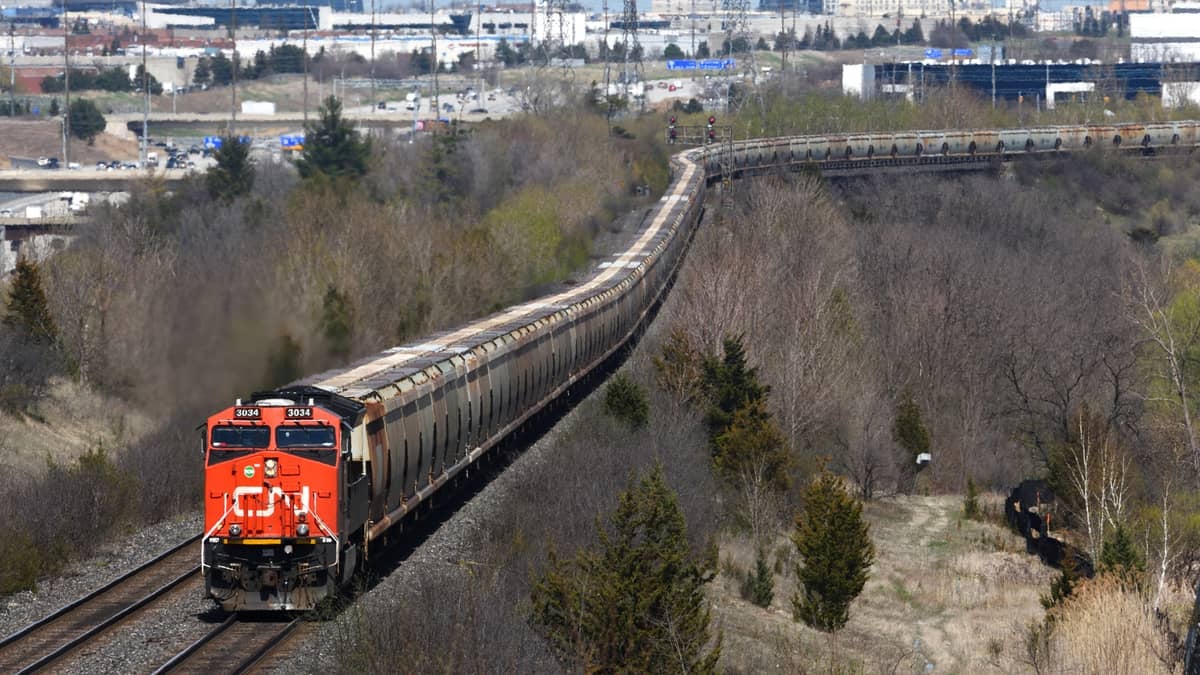 A photograph of a train hauling railcars. There are city buildings in the distance.