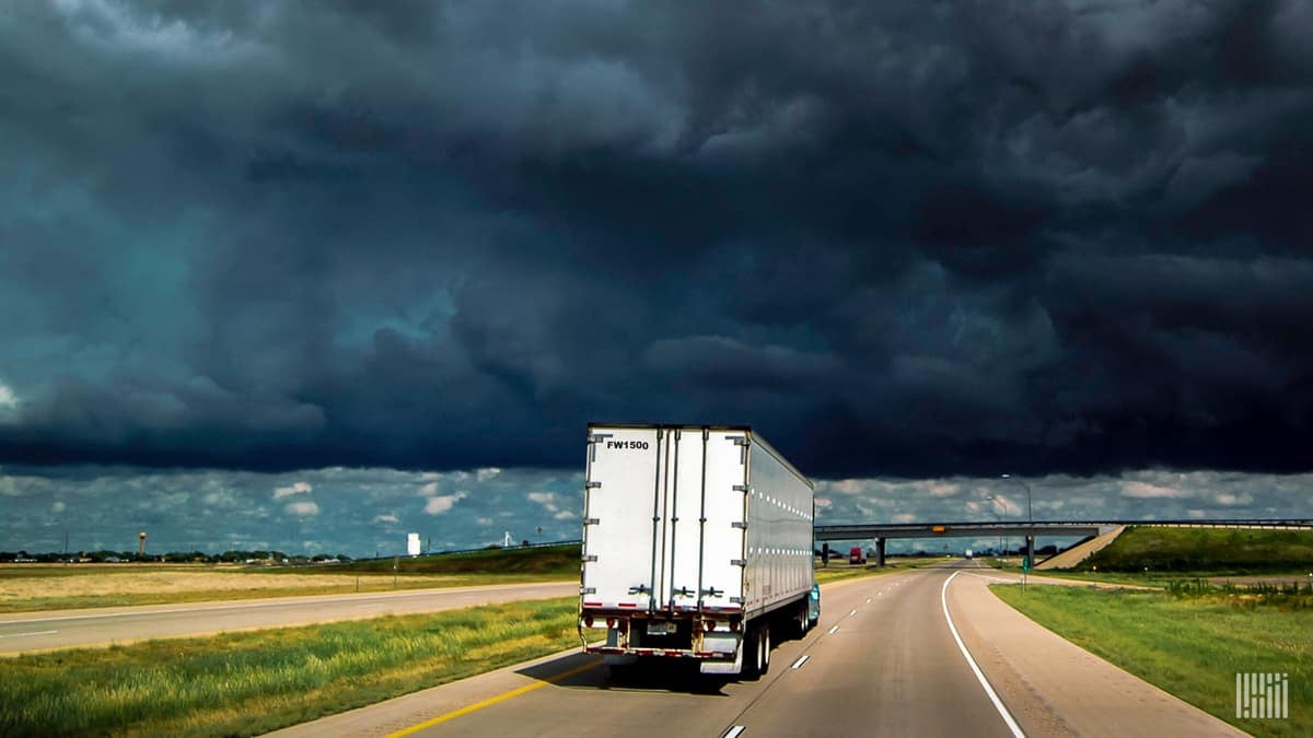 Tractor-trailer heading down highway with storm cloud ahead.