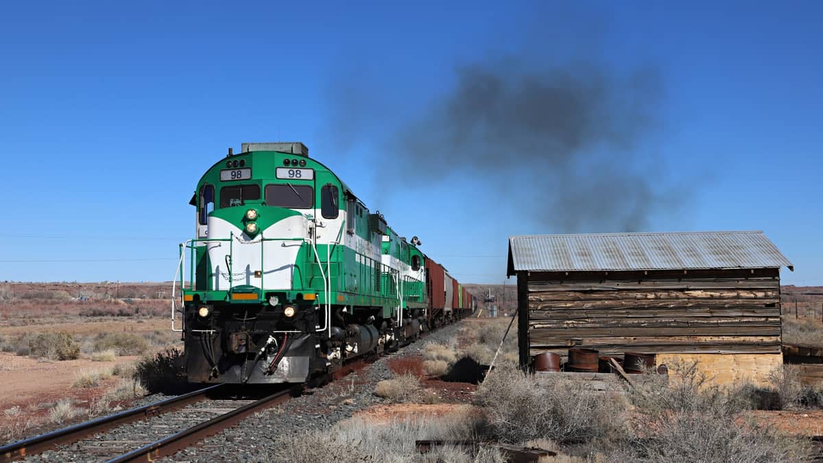 A photograph of a train passing by an old wooden barn in the middle of a field.