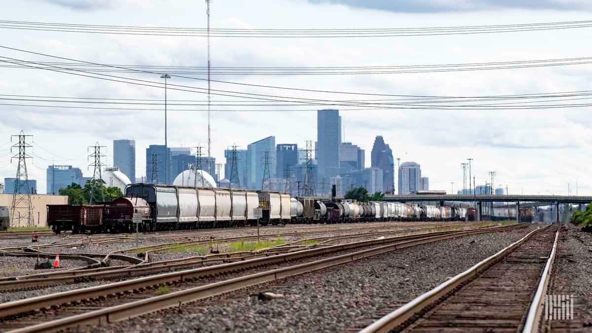 A photograph of a rail yard with train tracks and a train. There is a city with skyscrapers far in the distance behind the train.