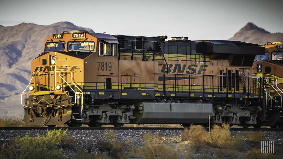 A photograph of a train traveling through a desert field. There is a mountain range in the distance behind the train.