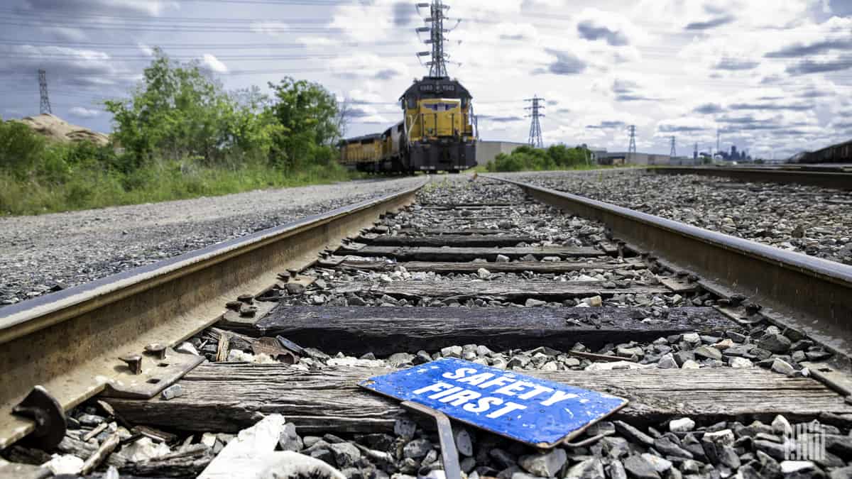 A photograph of a rail track close up. On the track is a paper sign that says, Safety First. In the distance is a freight train locomotive.