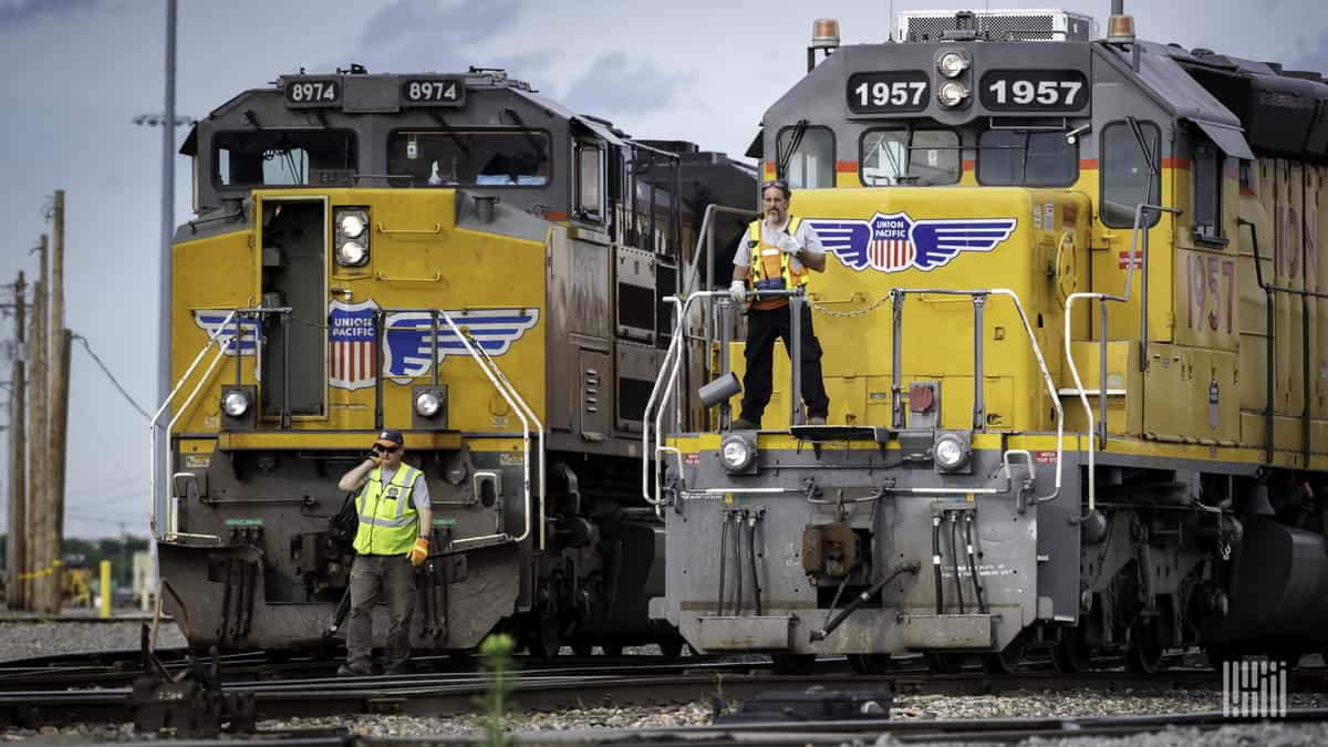 A photograph of two train locomotives. There are two men standing in front of each train.