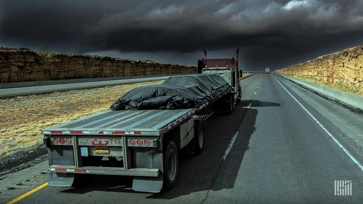 Flatbed truck heading down highway with dark storm cloud ahead.