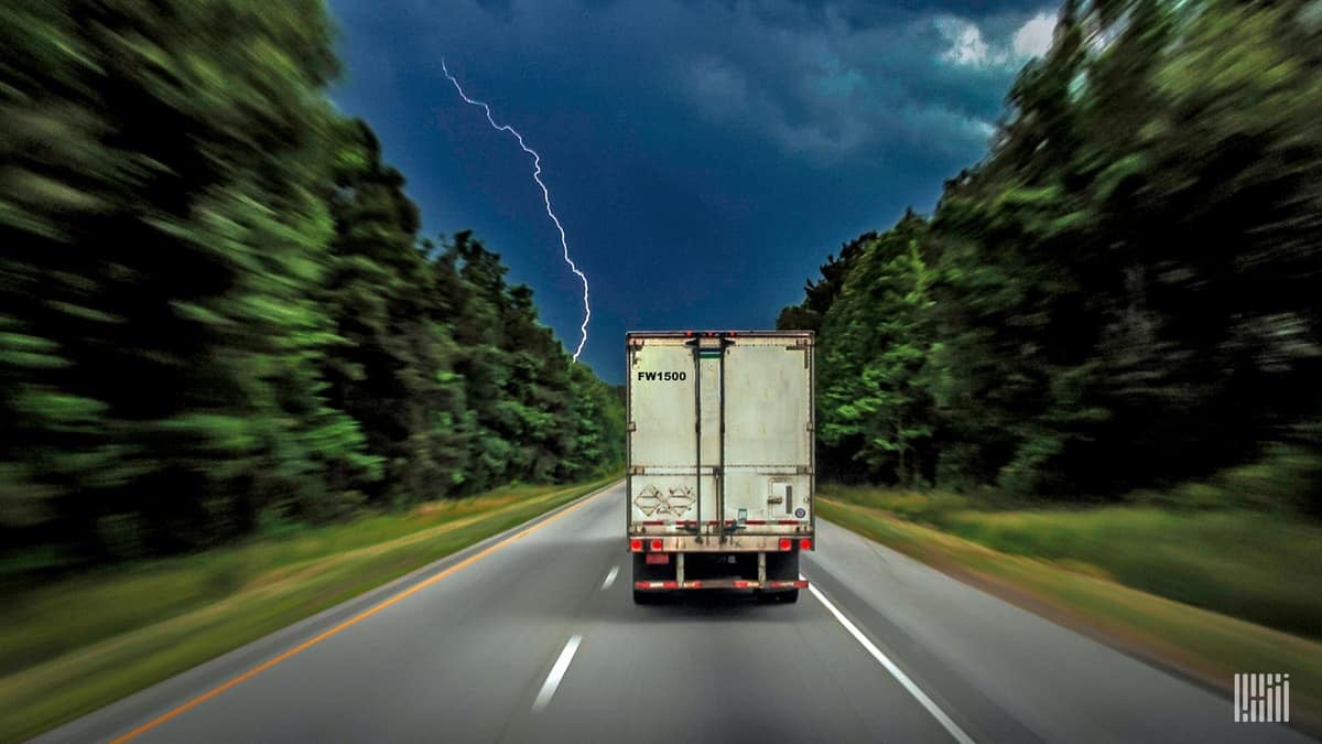 Tractor-trailer heading down highway with thunderstorm ahead.