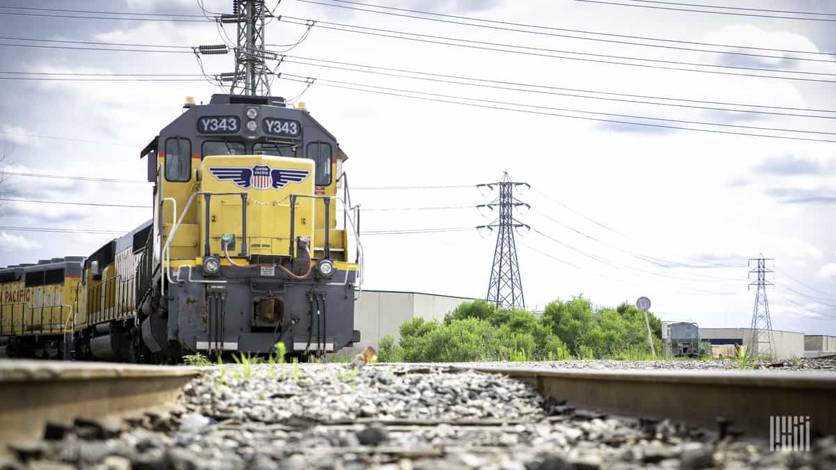 A photograph of a train in a rail yard. There are electricity lines above the train and behind it.