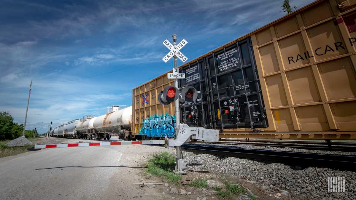 A photograph of a highway-rail grade crossing. There is a train with boxcars and tank cars crossing the intersection.