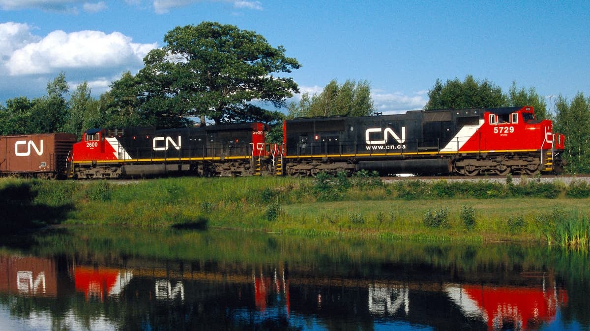 A photograph of a train traveling in a grassy field. A reflection of the train is on the water of a nearby pond.