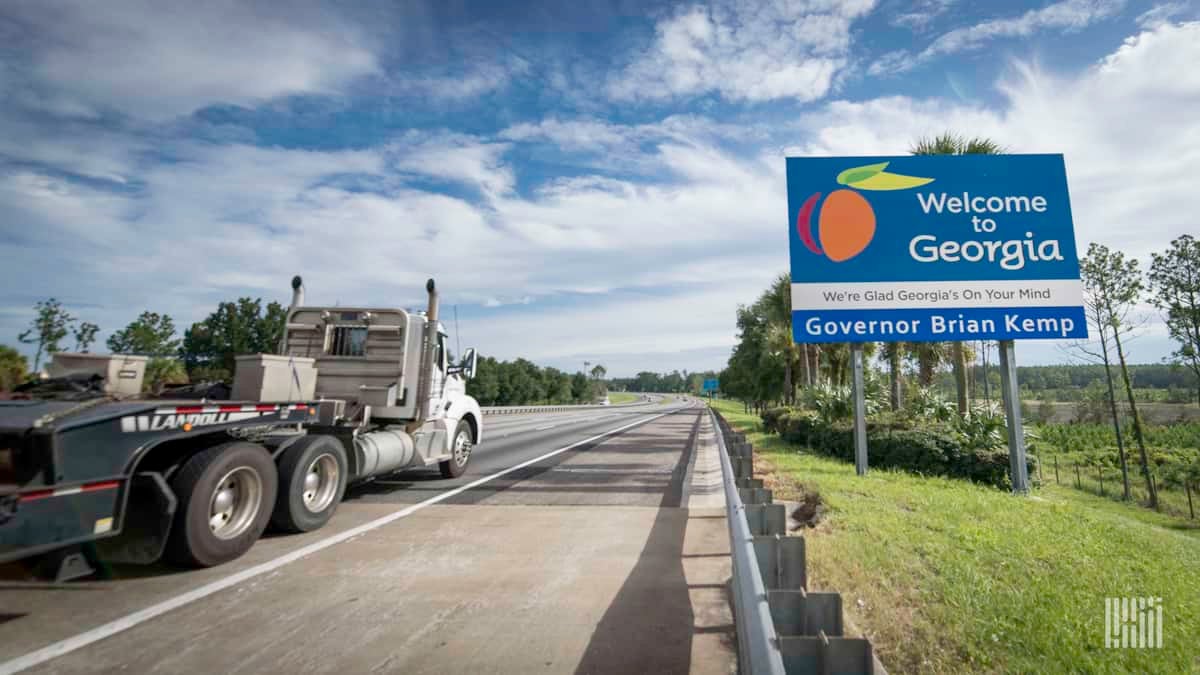 A flatbed commercial truck passes a sign that reads "Welcome to Gerogia"