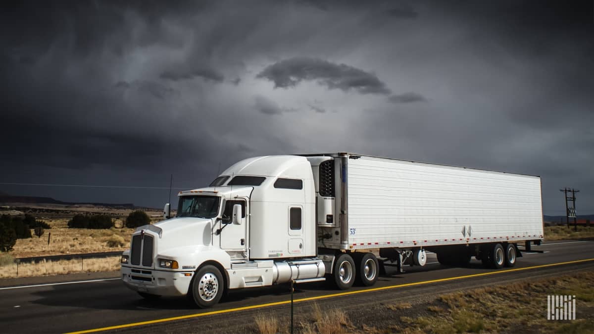 Tractor-trailer heading down highway with dark storm cloud in background.