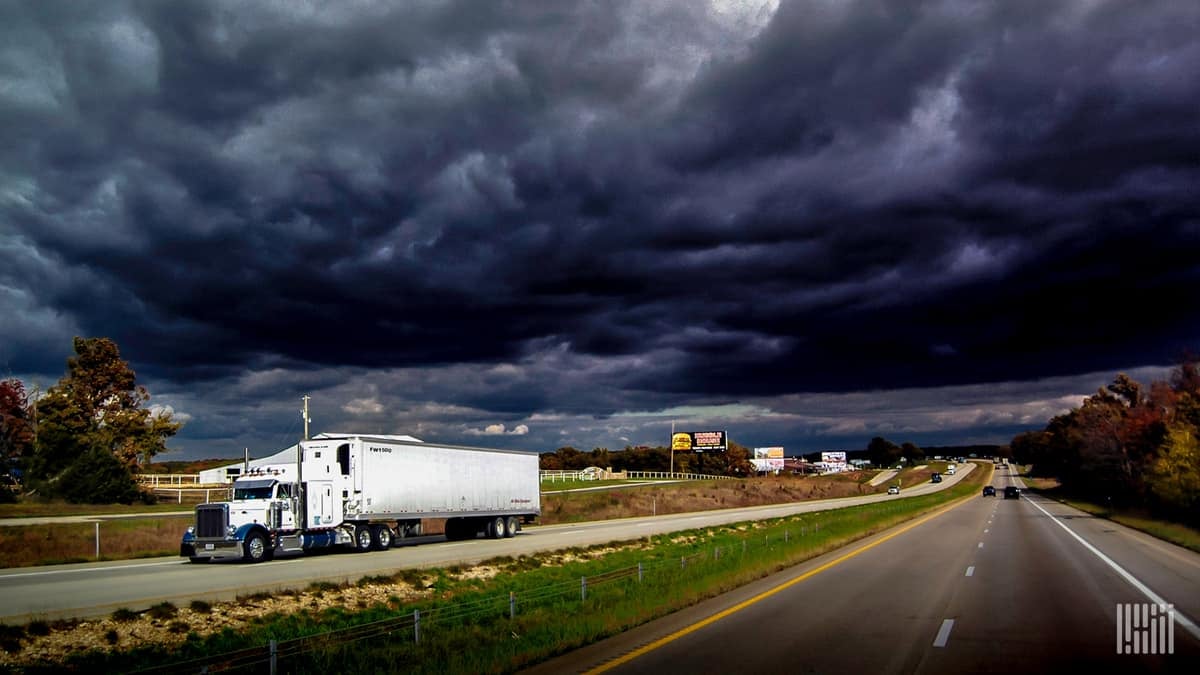 Tractor-trailer heading down highway with dark storm cloud across the sky.