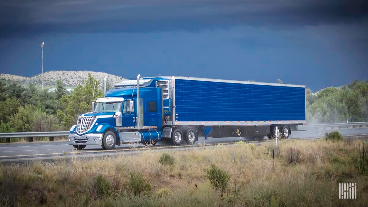 Tractor-trailer heading down highway with dark storm cloud in the background.