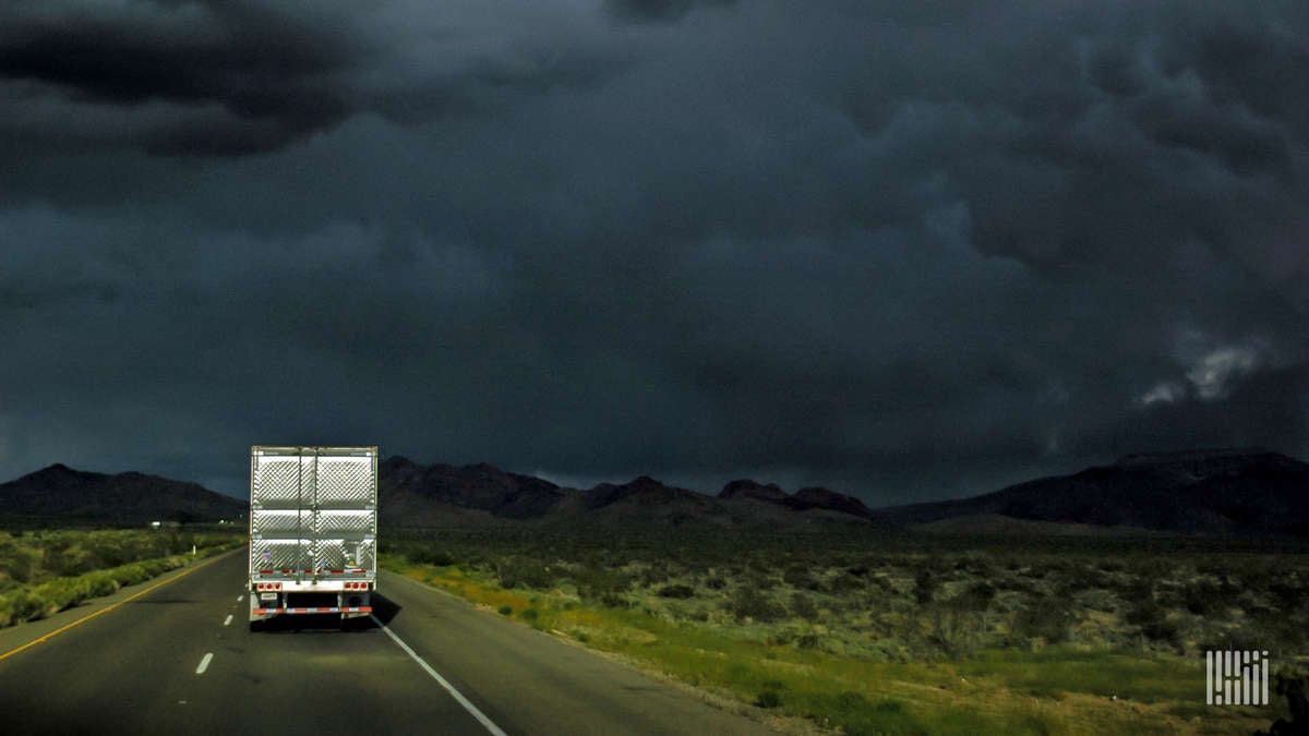 Tractor-trailer heading down highway with very dark storm cloud ahead.