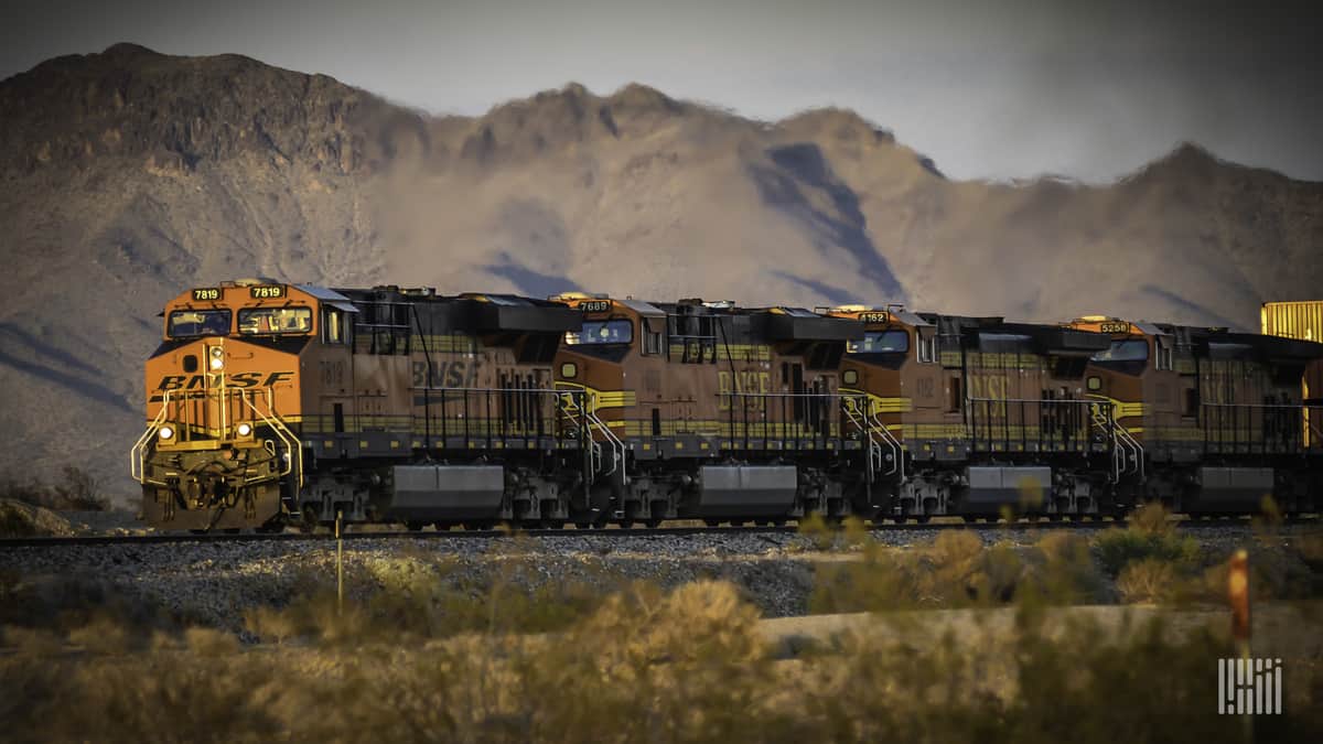 A photograph of a BNSF train traveling through an open field of dry land. A mountain range is in the background.