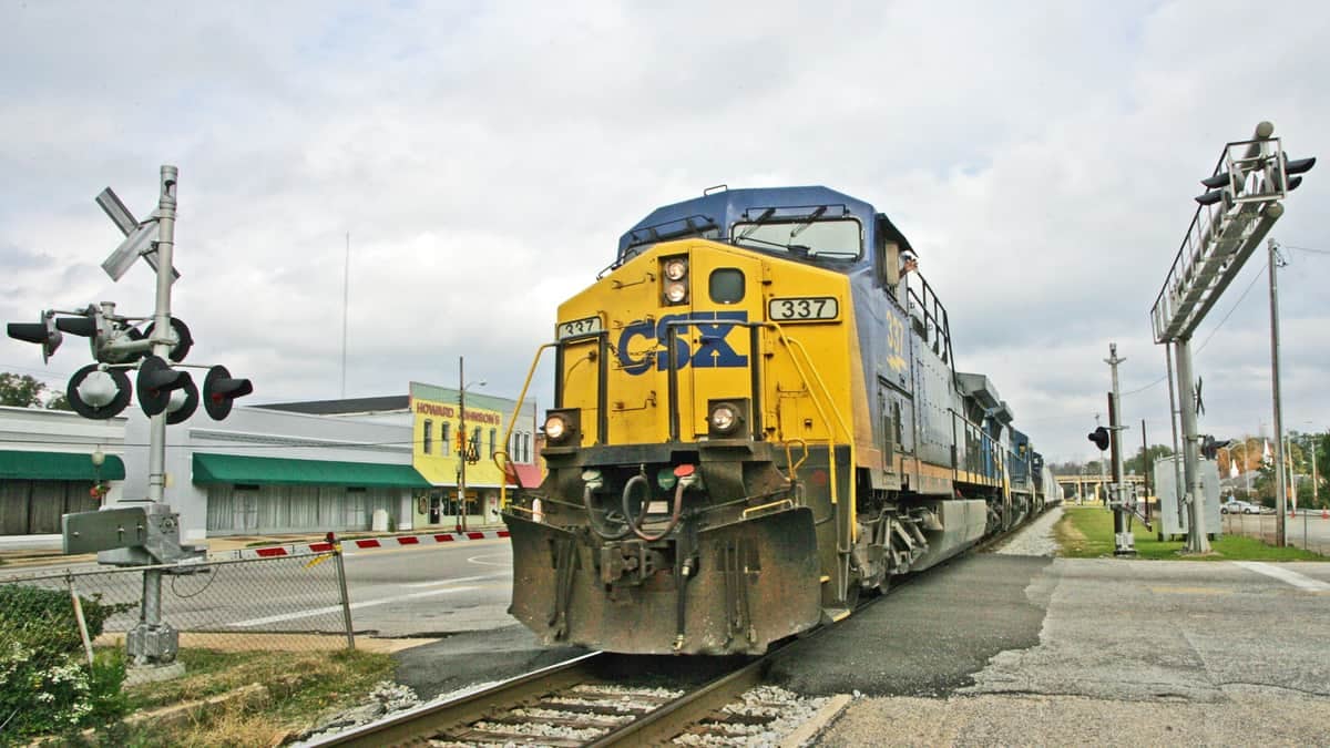 A photograph of a CSX train at a rail crossing. There are town buildings nearby the train tracks.