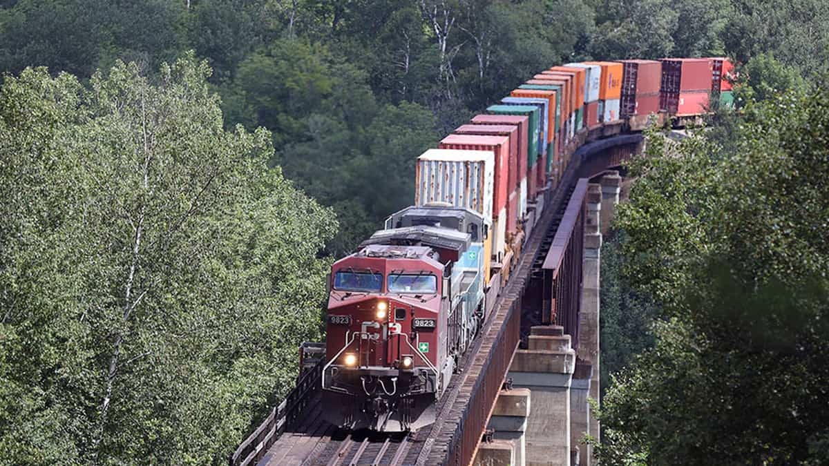 A photograph of a Canadian Pacific train carrying containers. The train is traveling through a forest.