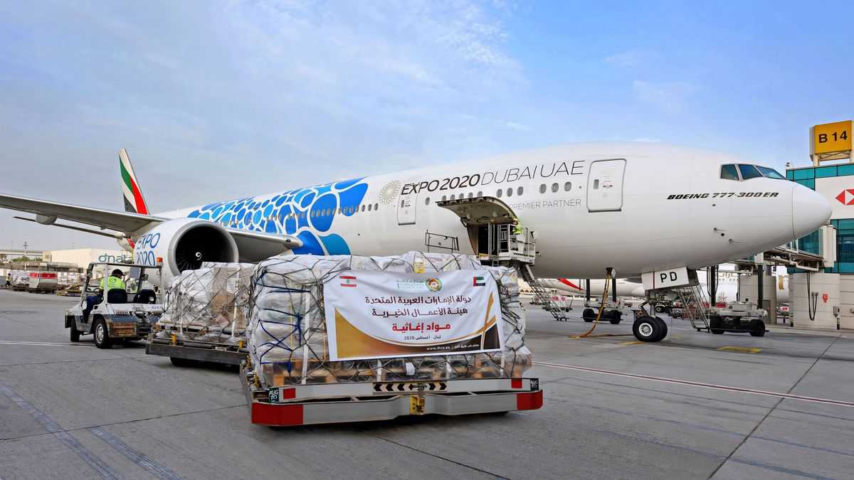 A pallet of emergency supplies with a sign in Arabic sitting on the tarmac next to an Emirates plane on a sunny day. Emirates is flying supplies to Beirut to help the city recover from the explosion.