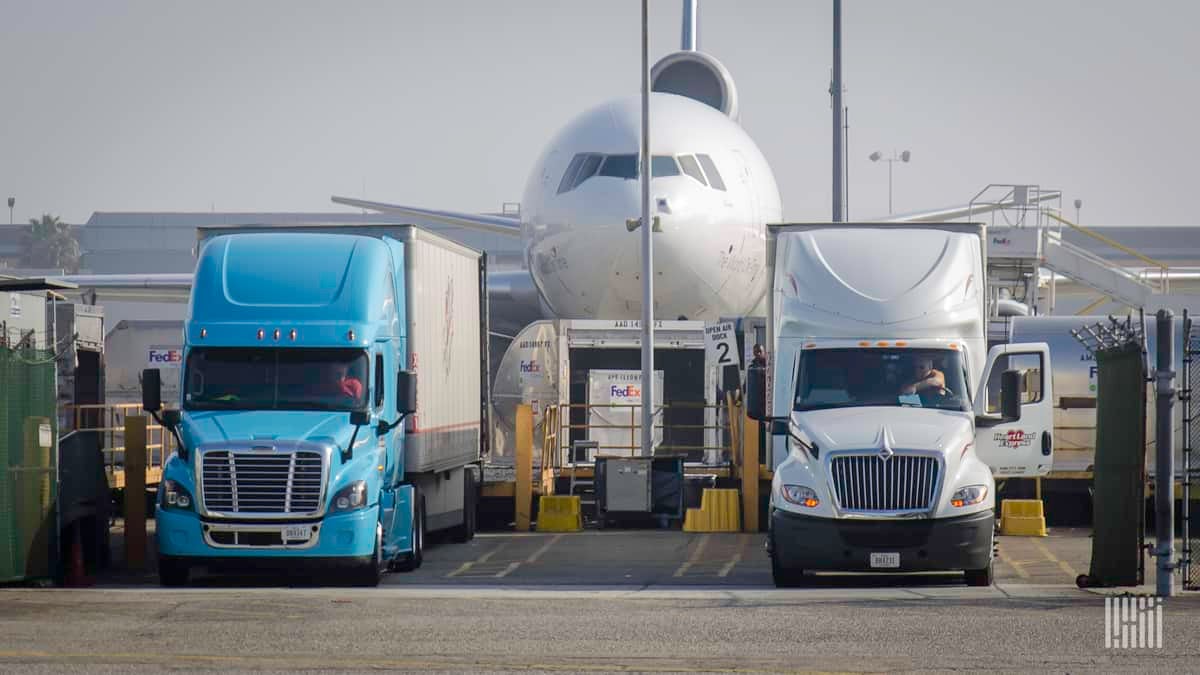 A white FedEx cargo jet flanked by large trucks. The air cargo market is volatile this summer and prices are heading up on key lanes.