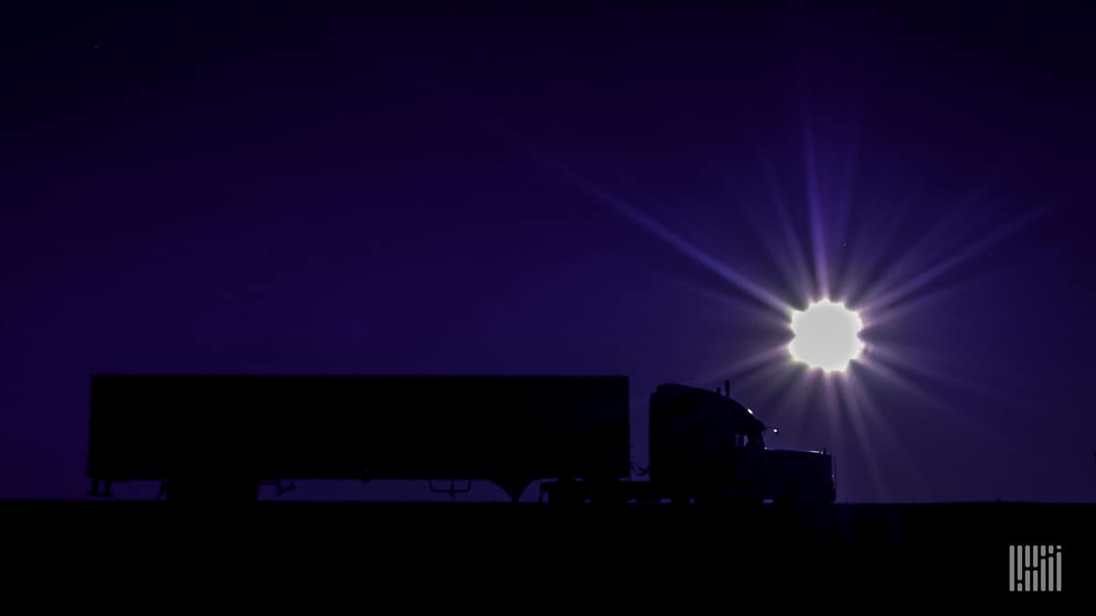 Tractor-trailer heading down highway with bright sun on the dusk/dawn horizon.