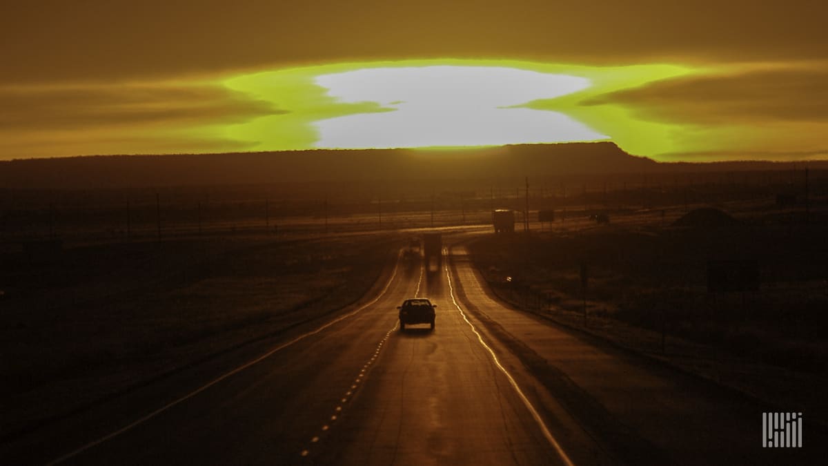 Tractor-trailers and cars heading down desert highway with bright sun on the horizon.