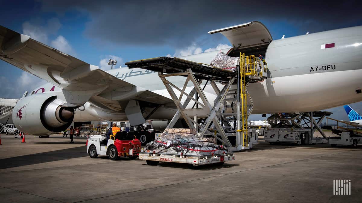 A big gray plane getting loaded with cargo pallets from side door with hydraulic lift on a sunny day.